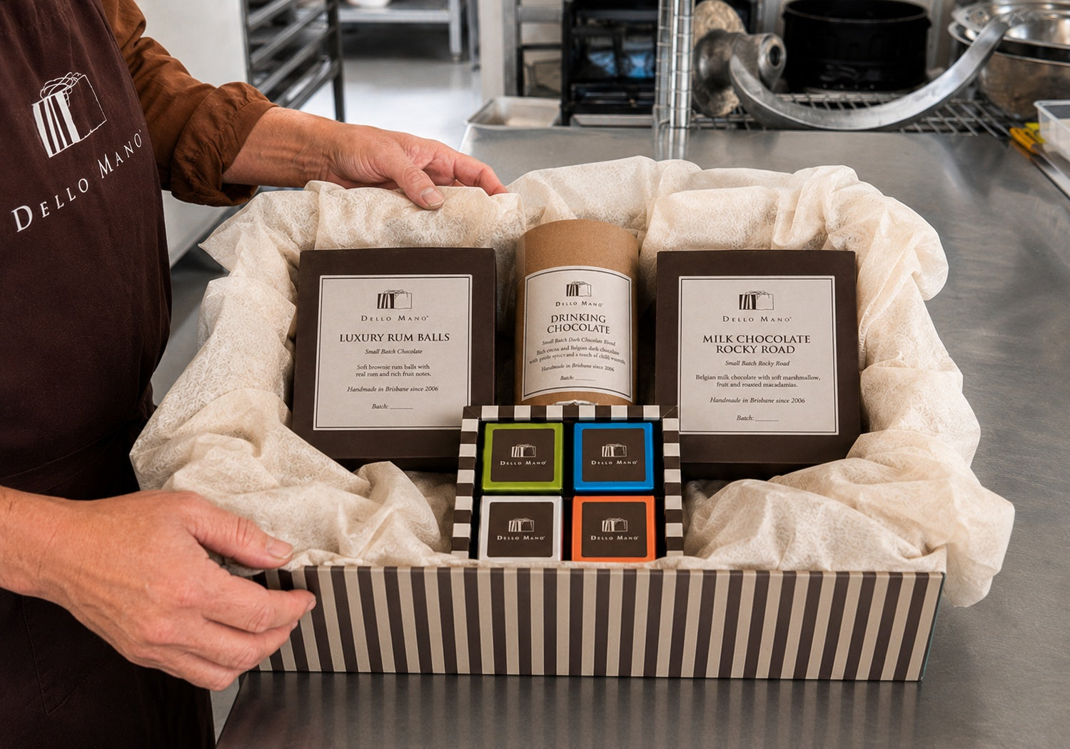 Dello Mano Chocolate Discovery Hamper being packed in the West End kitchen with a four-piece brownie gift box, Dark Drinking Chocolate, Luxury Rum Balls and Milk Chocolate Rocky Road arranged in a deep striped hamper box with cream tissue.