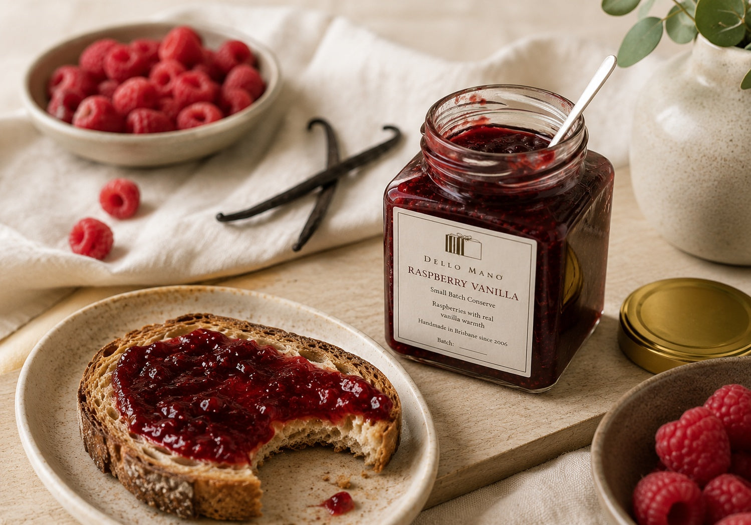 Open jar of Dello Mano Raspberry Vanilla Jam served with artisan toast, fresh raspberries and vanilla beans in a calm natural light breakfast setting.