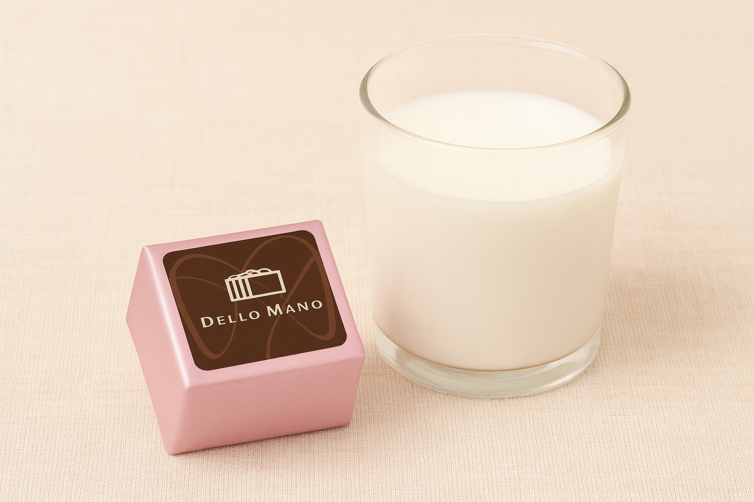 Dello Mano Peanut Butter Brownie Cube wrapped in pale pink foil with branded label, styled on a beige linen background beside a clear glass of milk.