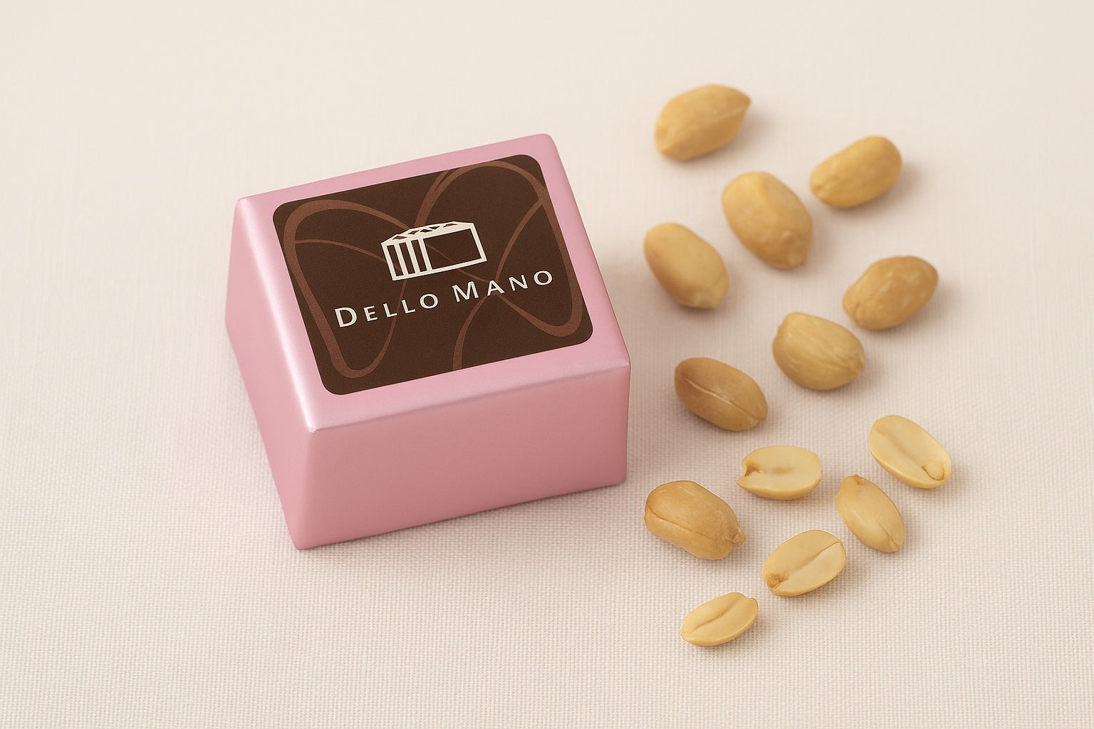 Dello Mano Peanut Butter Brownie Cube wrapped in pale pink foil with branded label, styled on a beige linen background with roasted peanuts scattered around.
