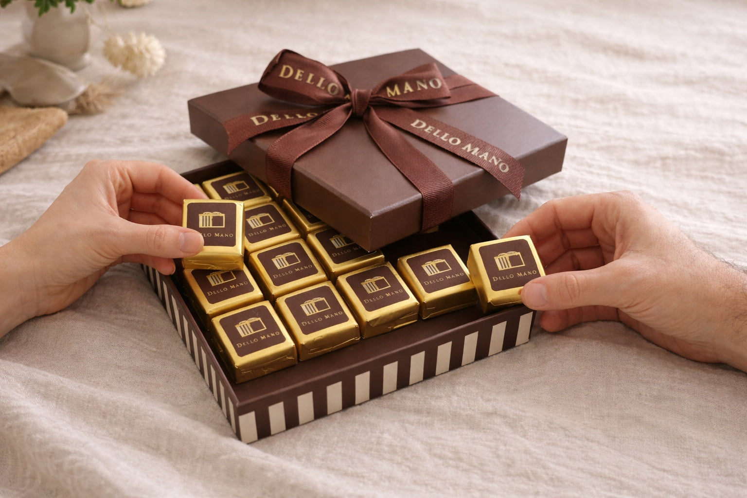 Open brownie gift box with handmade chocolate brownies in gold foil being shared by two hands on a soft linen background
