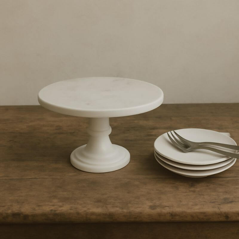 White marble cake stand on a timber surface with cake plates and cake forks.