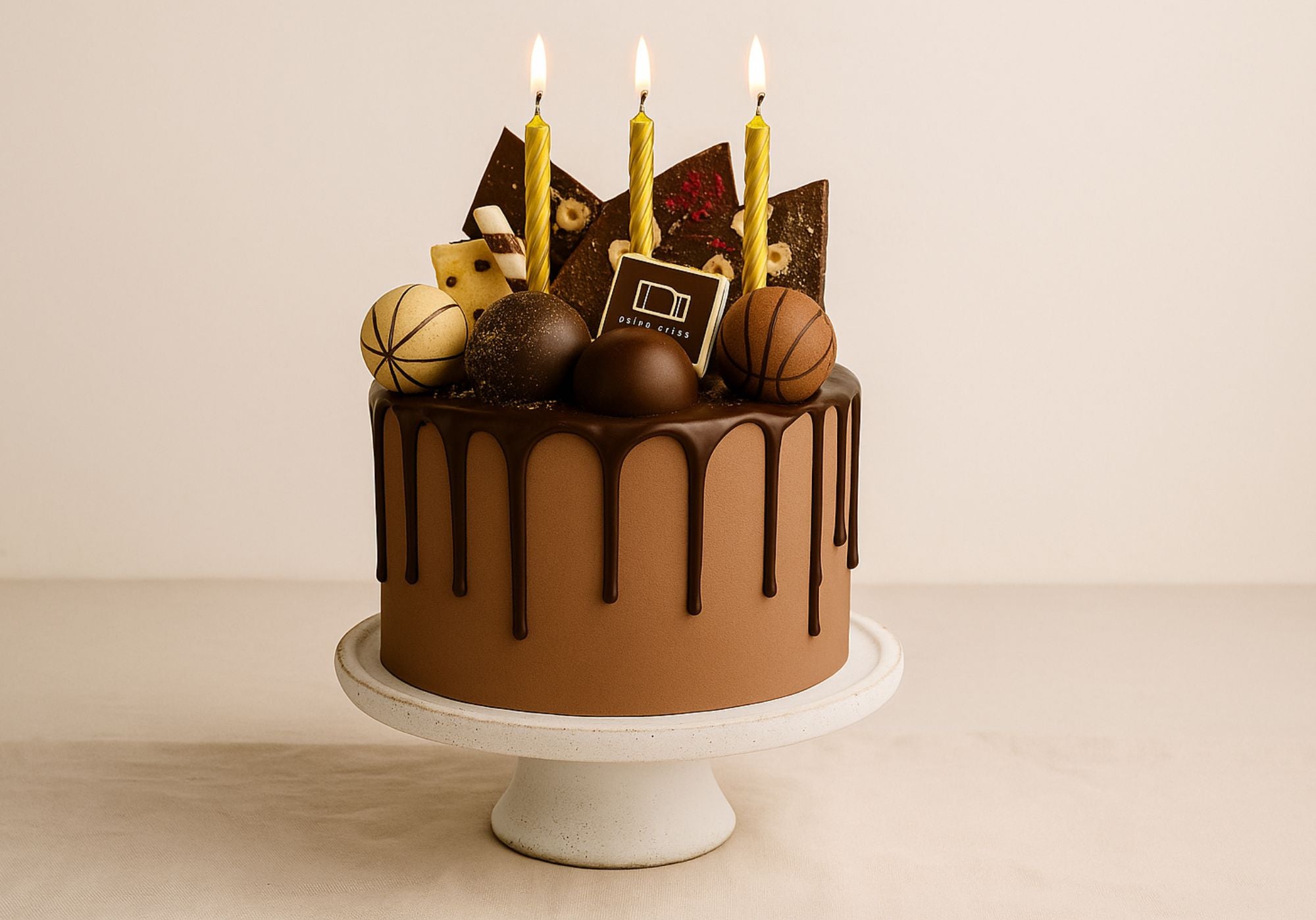 Chocolate birthday cake with golden candles, chocolate drip, truffles, and decorative bark, displayed on a ceramic stand.