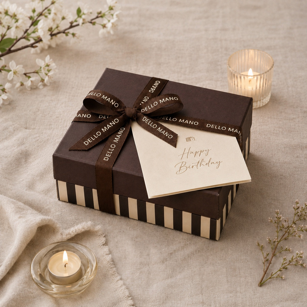 Dello Mano birthday gift box tied with signature ribbon, accompanied by a Happy Birthday card and candles on soft linen.