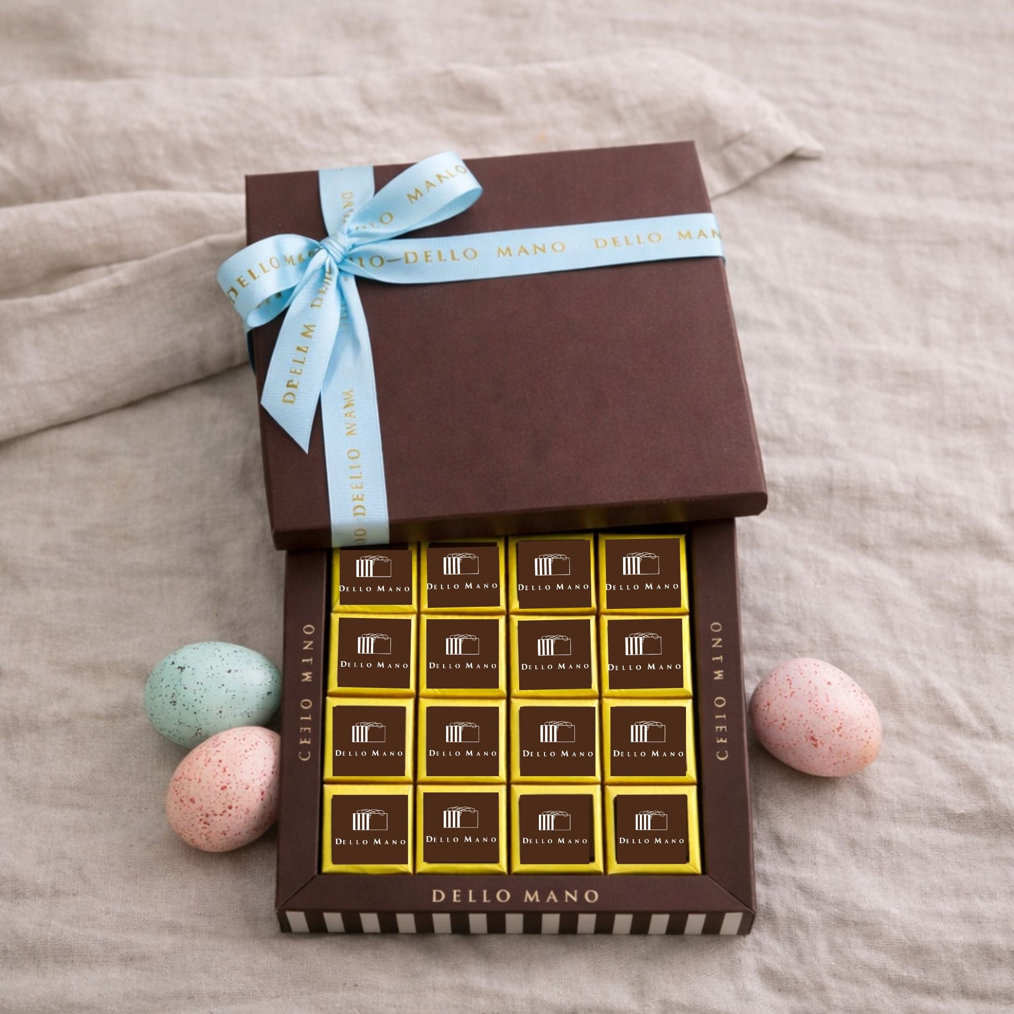 Dello Mano Classic Luxury Brownie gift box with pastel blue ribbon and Easter eggs on natural linen background.