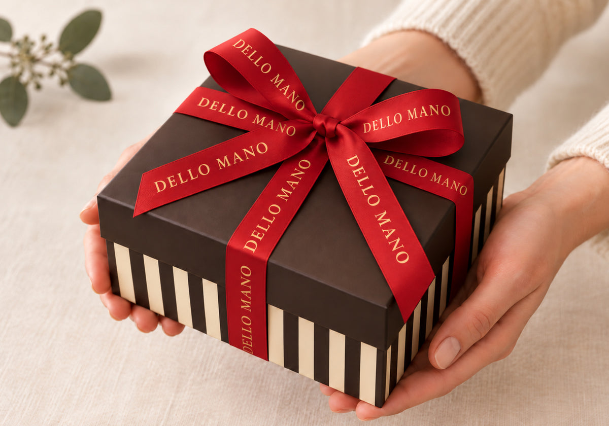Hands holding a striped Dello Mano Christmas gift box tied with a red branded ribbon.