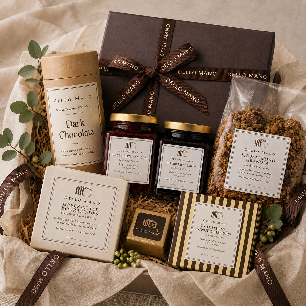 Dello Mano gift hamper with brownies, biscuits, granola, conserves and drinking chocolate arranged on soft neutral linen.