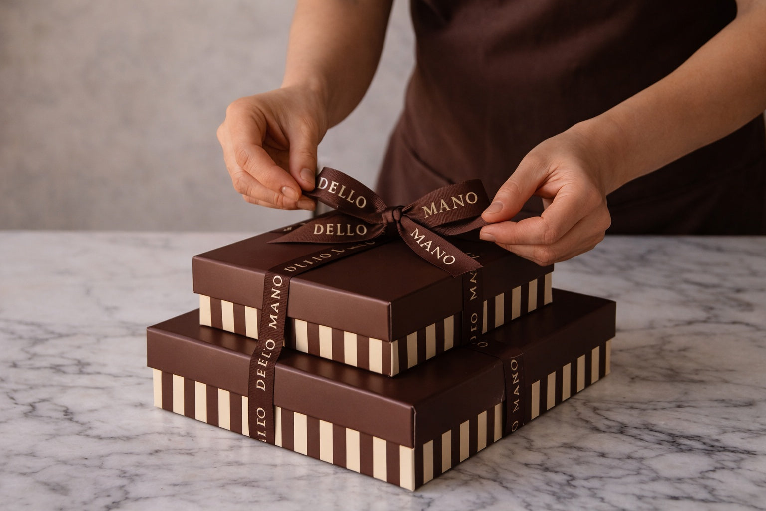 Hand tying ribbon on Dello Mano gift boxes on marble surface, showcasing handmade chocolate gifting