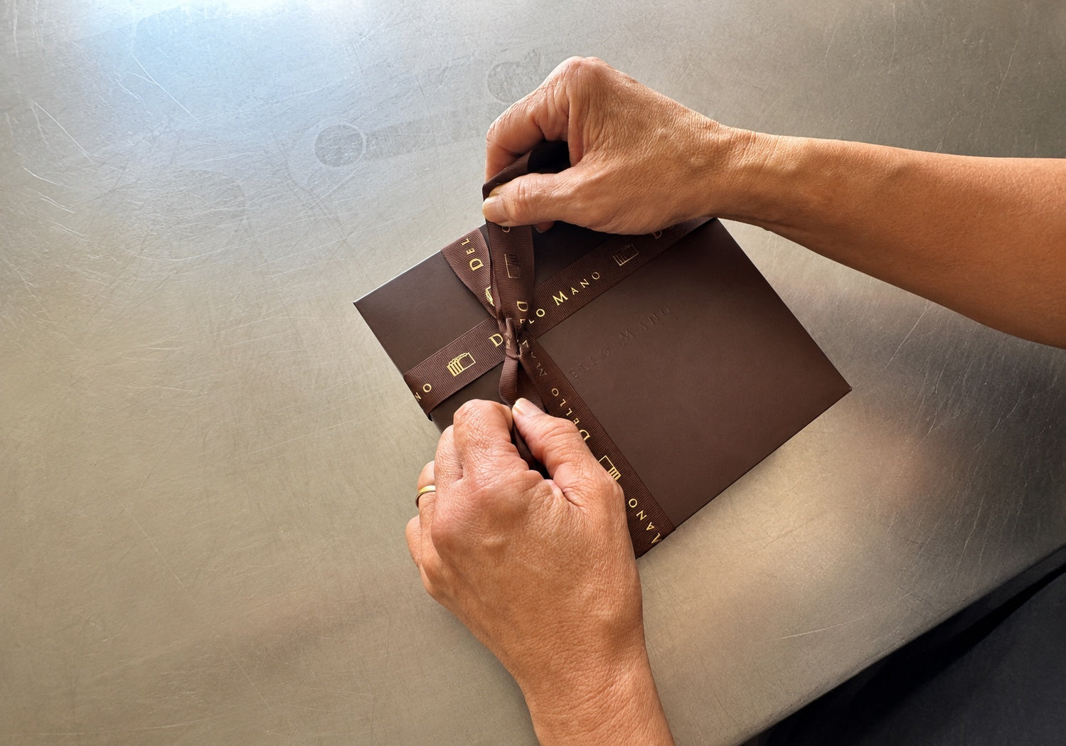 Hands tying Dello Mano ribbon around a handmade gift box on a stainless-steel kitchen bench.