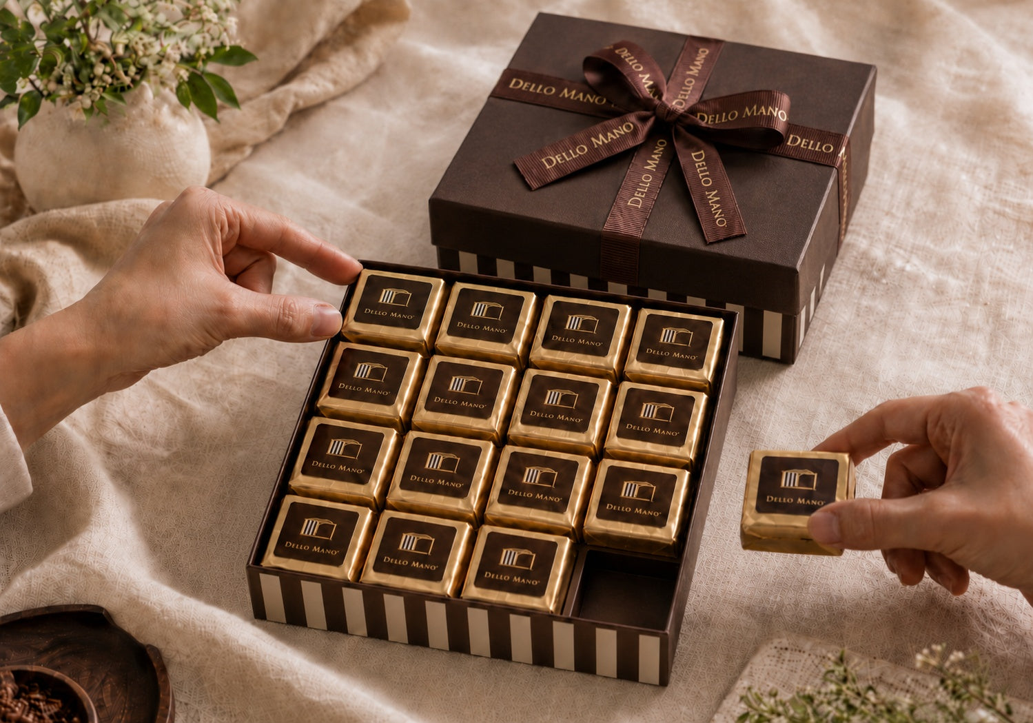 Open Dello Mano 16-piece brownie gift box with individually wrapped gold cubes, one piece removed, and a ribboned box behind.