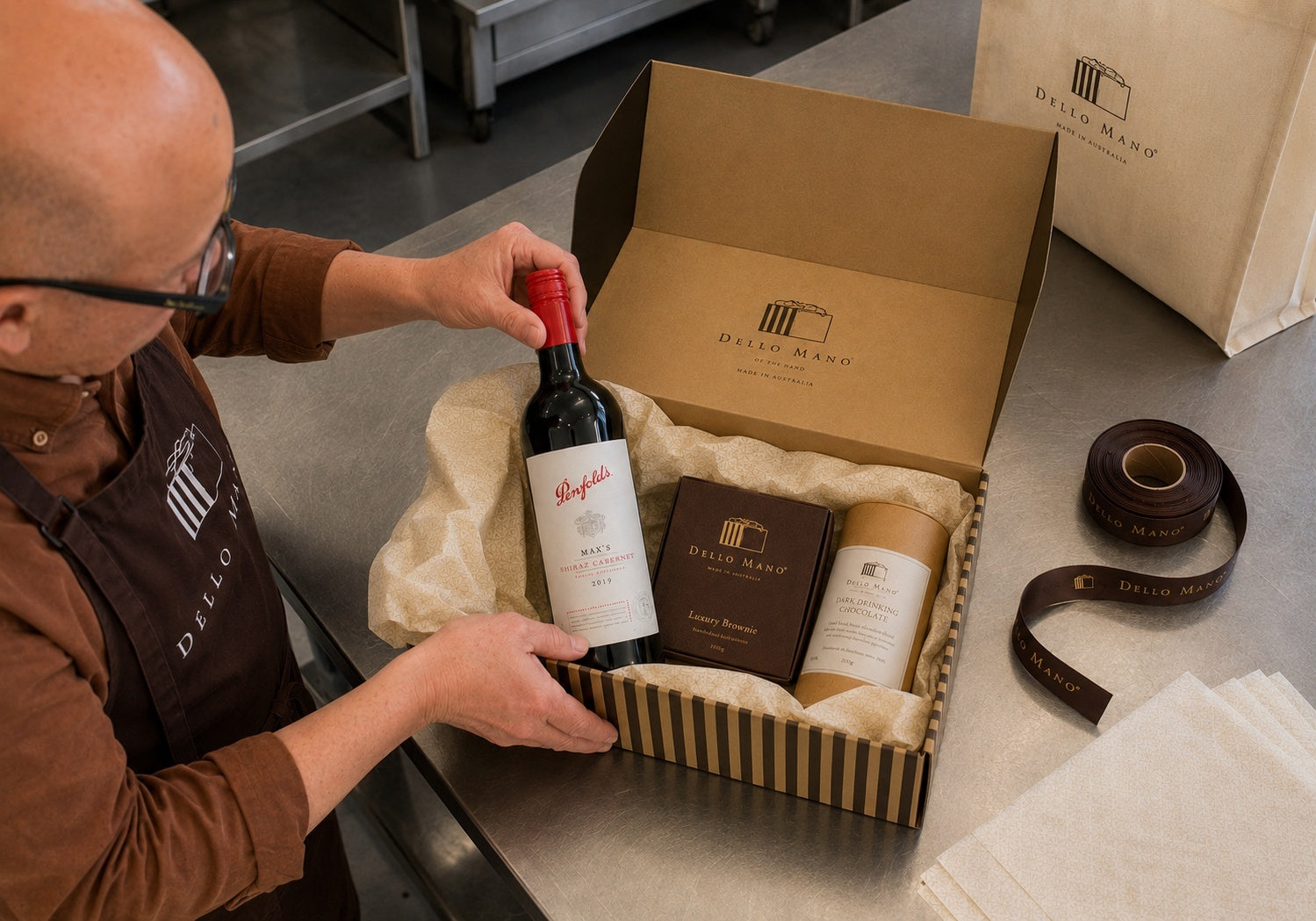 Bien placing a bottle of red wine into a striped Dello Mano hamper box with cream tissue, brownies and drinking chocolate on a stainless-steel bench in the West End kitchen.