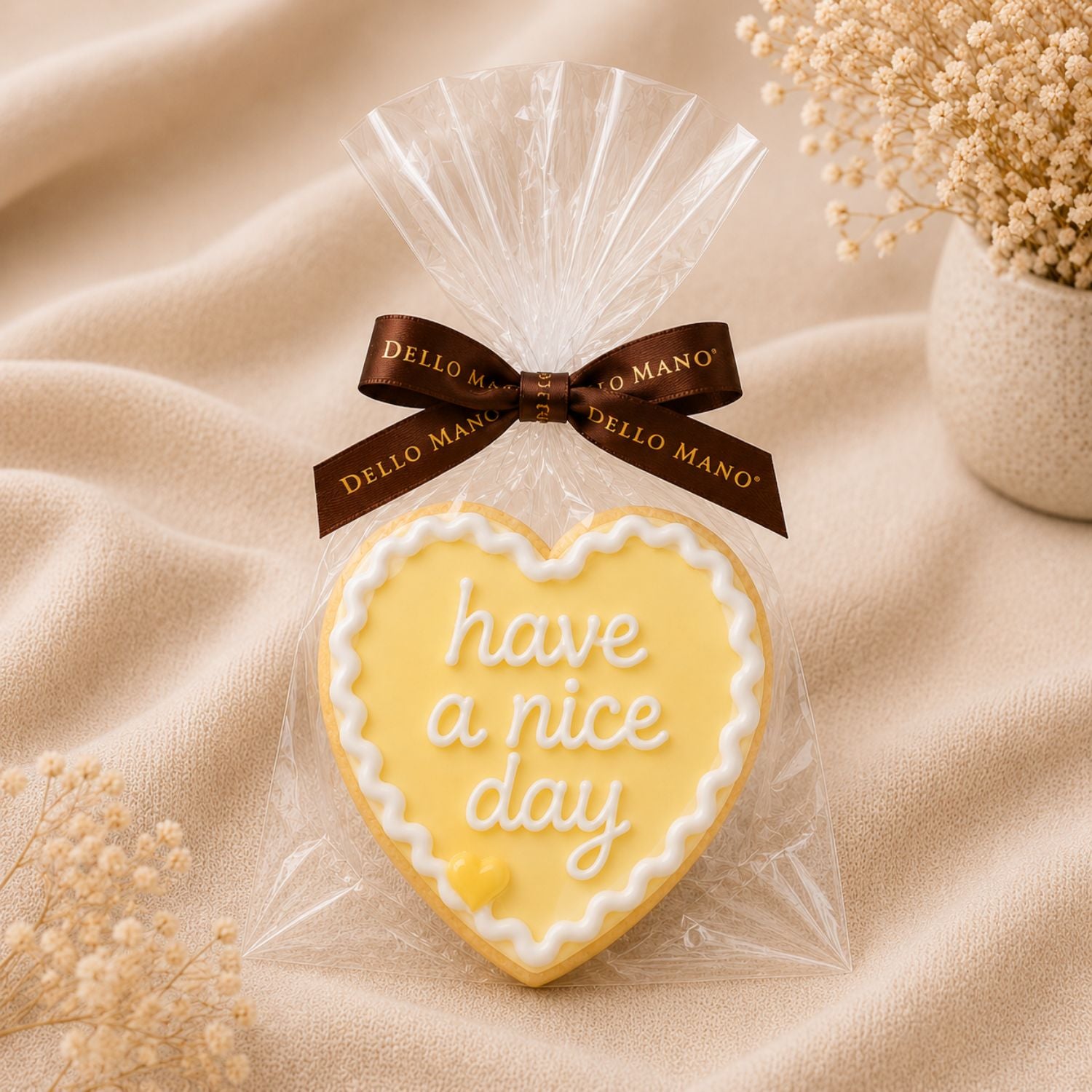 A lemon coloured sugar cookie saying "have a nice day"wrapped in clear cello with brown Dello Mano ribbon, styled on soft neutral fabric with delicate dried flowers.
