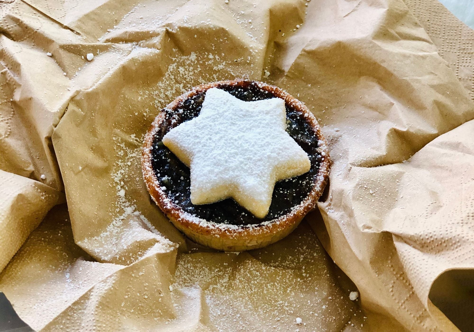 Handmade Christmas mince pie topped with a buttery shortbread star and dusted with icing sugar, presented on rustic brown paper.