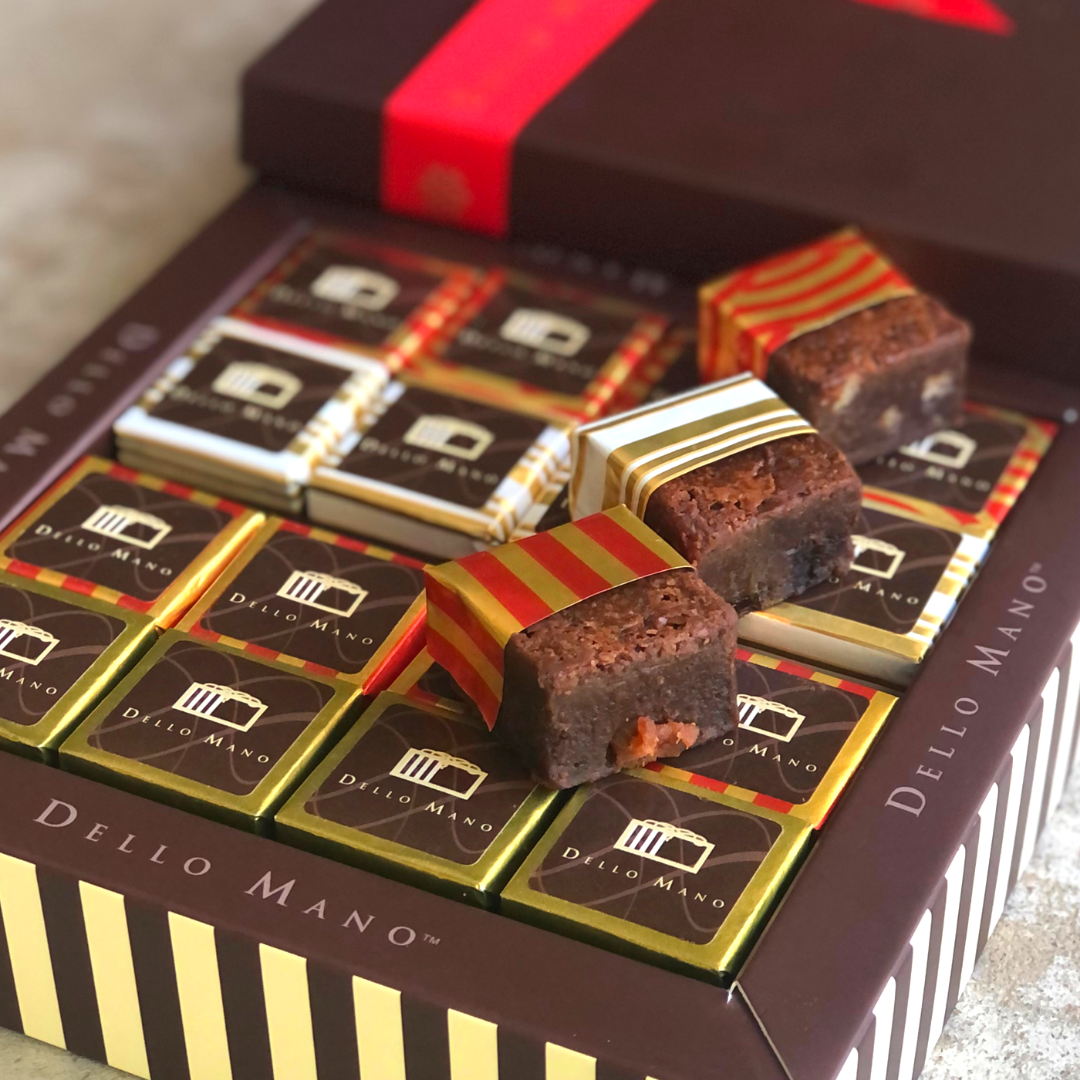 A brown chocolate gift box with stripes that says Dello Mano filled with Christmas brownies foil covered. Some brownies on top have foil open with brownie showing. 