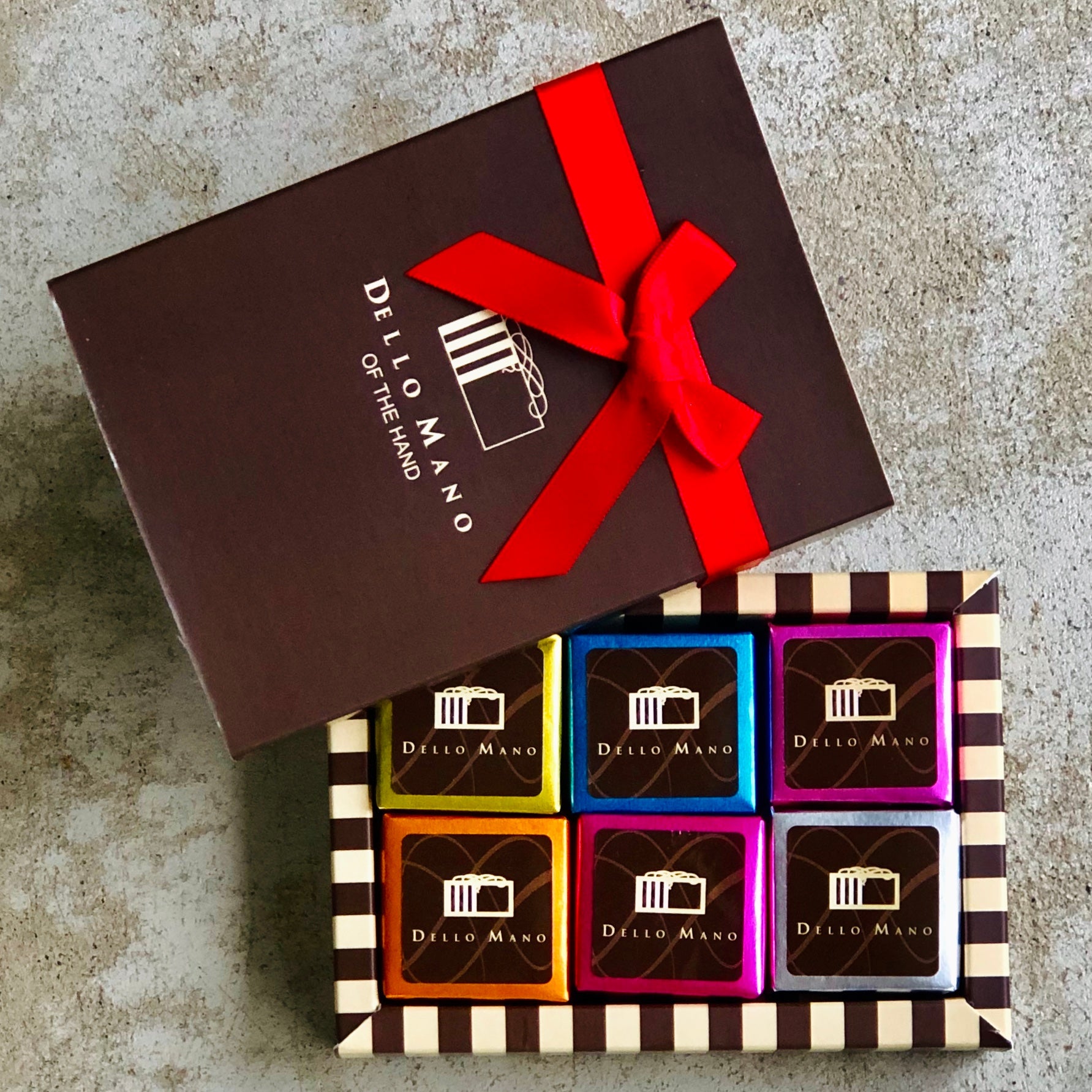 A small gift box of multi coloured brownies in a striped gift box. a lid to the box sits at the side with red ribbon and the words Dello Mano of the hand