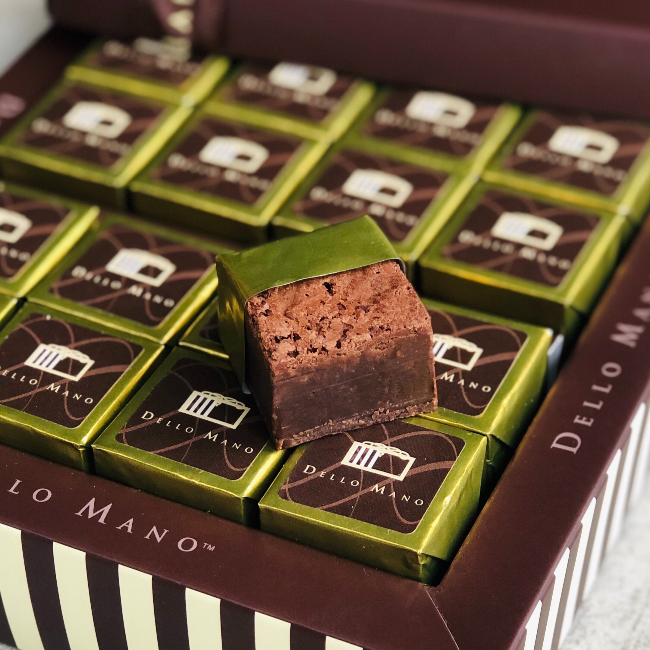 An open chocolate gift box of gold individually wrapped Brownie cubes. The box has words saying Dello Mano and there is an open foil brownie on top of the box.