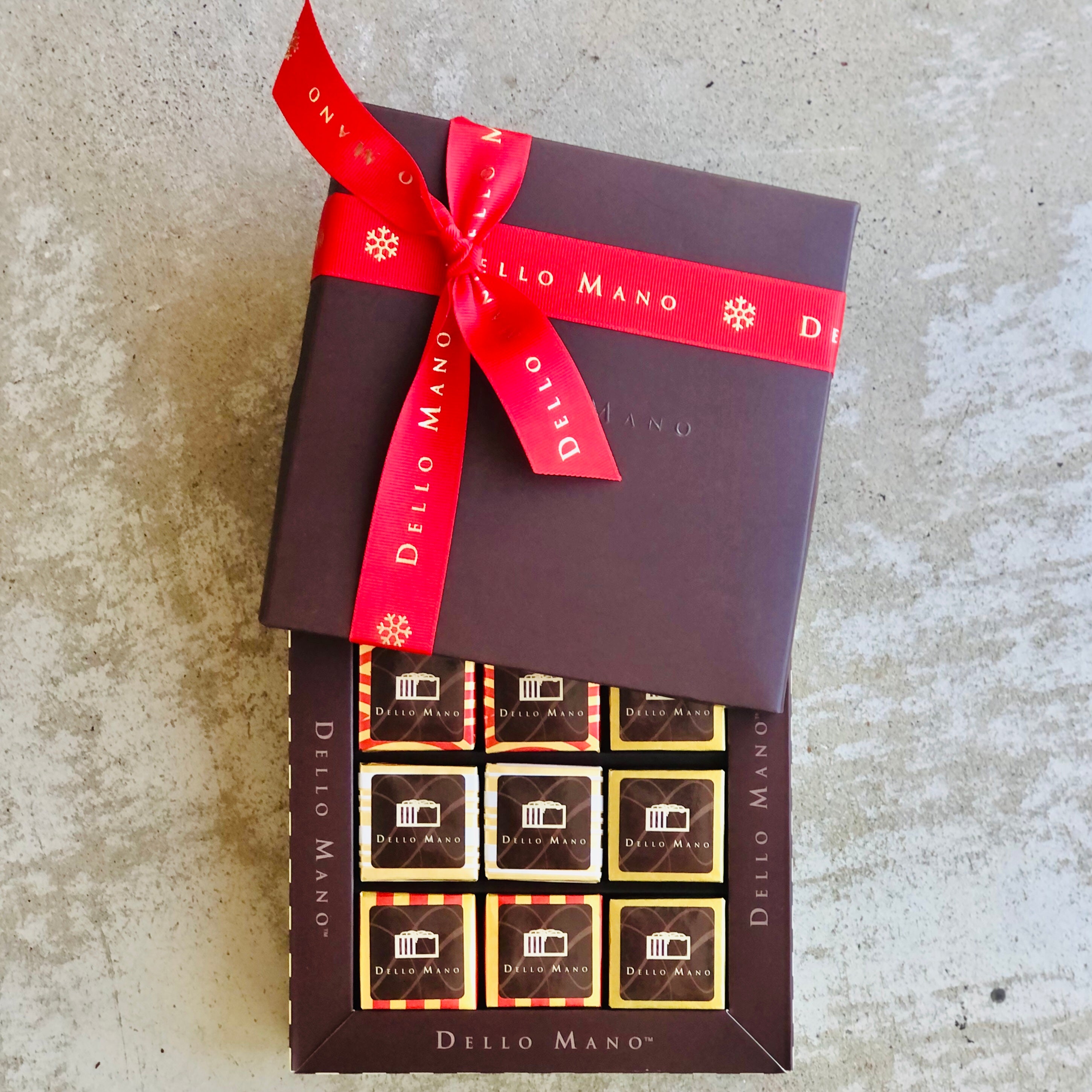 9 pieve gift box of Christmas brownies. The lid is open and has a red ribbon that says Dello Mano. Each brownie chocolate is individually wrapped. 