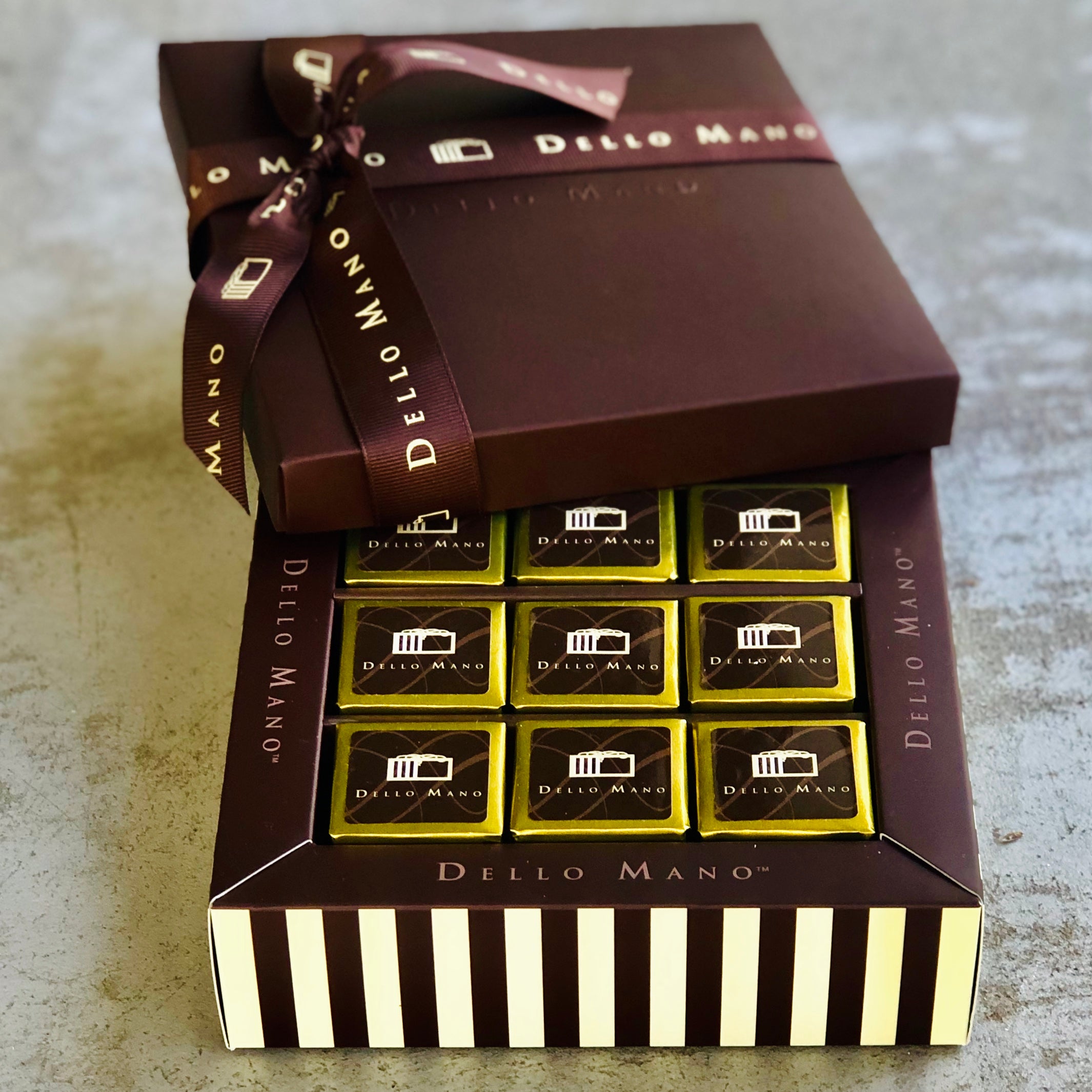 Nine individually foiled brownie cubes in a striped gift box with the words Dello Mano. The lid sits open with a brown ribbon tie that also says Dello Mano