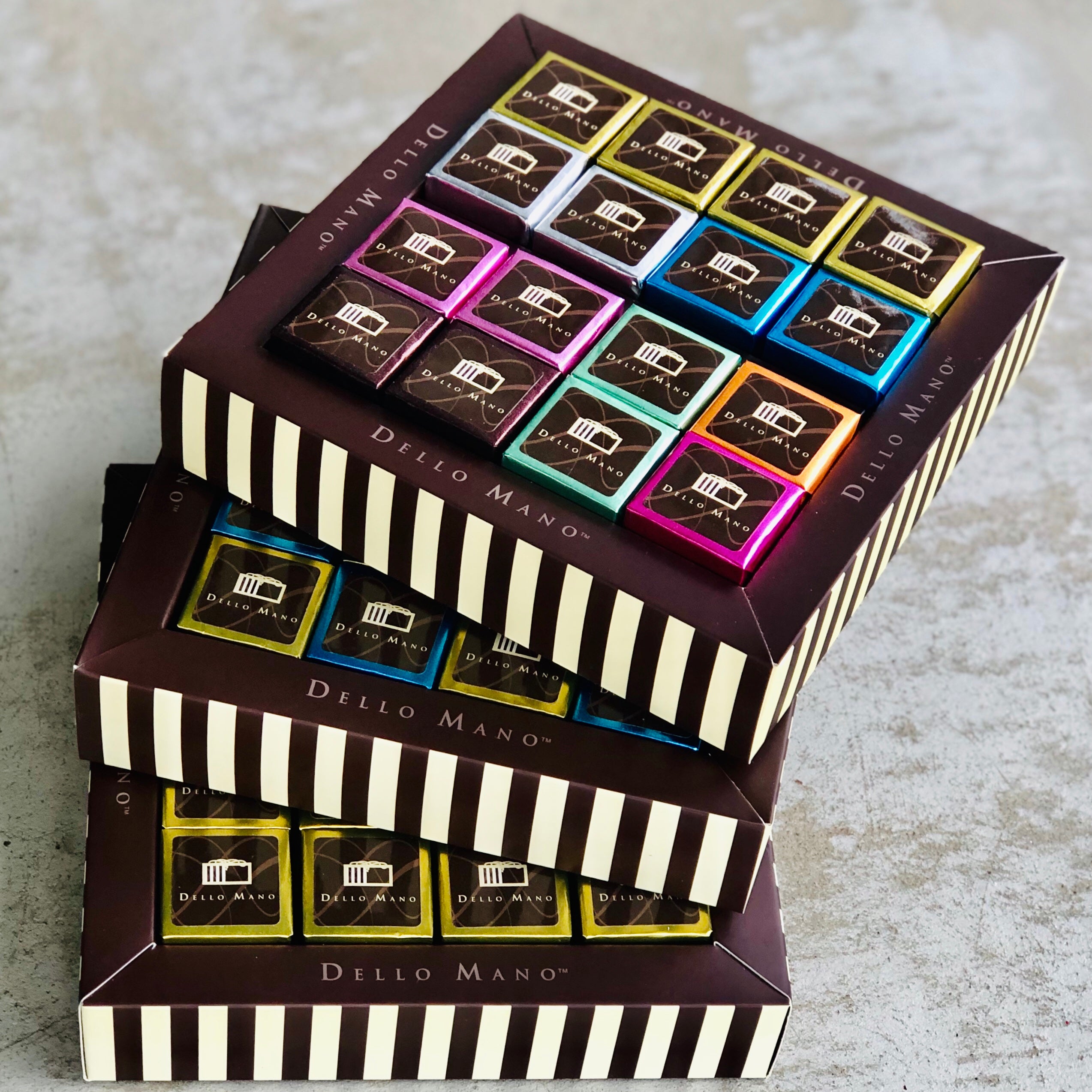 Three brown stiped boxes stacked on top of each other with individually foiled brownies. The chocolate gift box says Dello Mano