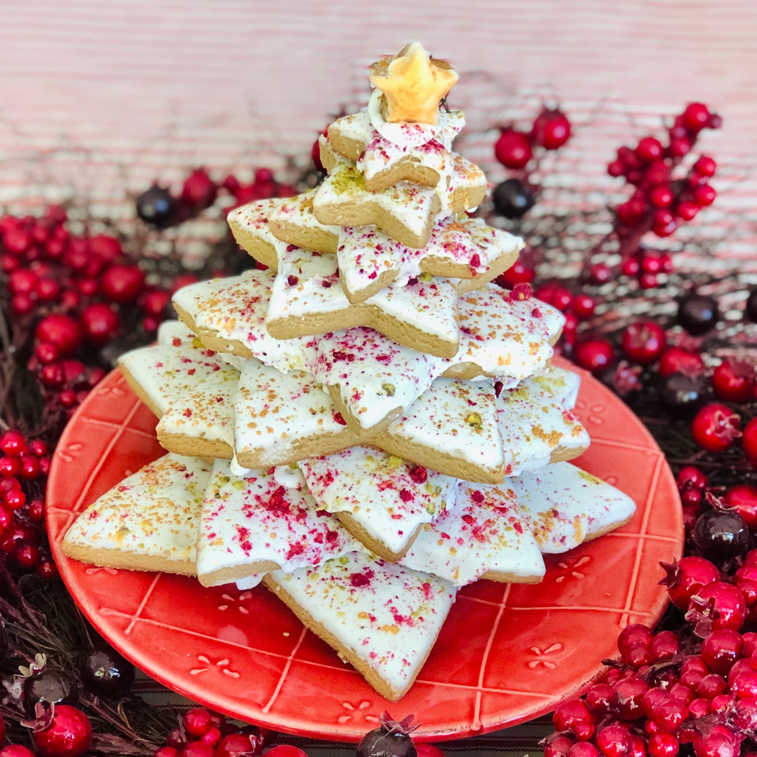 A gingerbread Christmas tree with gold and red sprinkles on a red plate with berries surrounding.