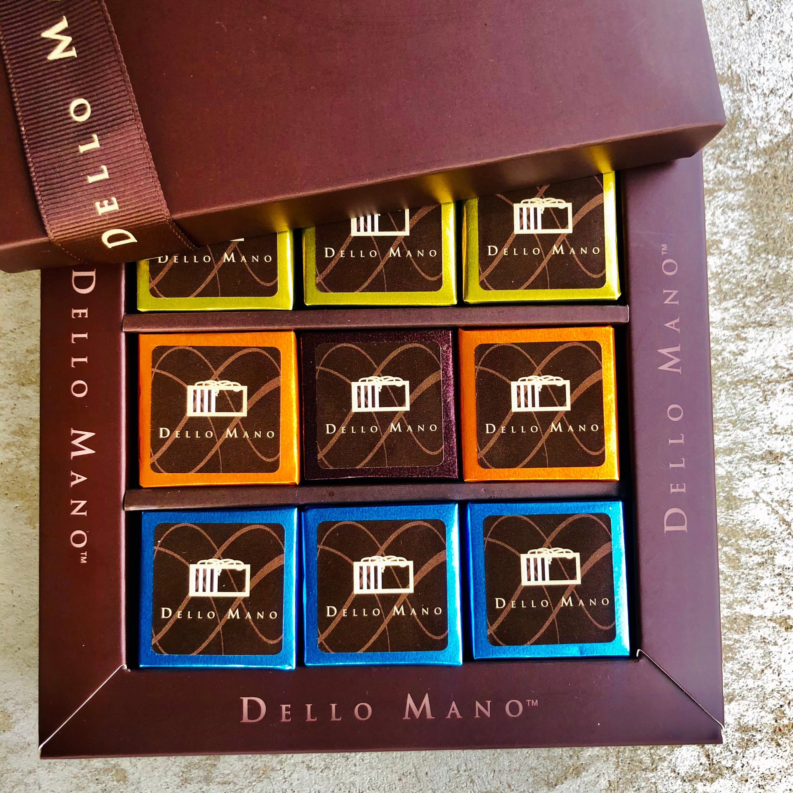 NIne brownie cubes sitting in an open chocolate box. The box says Dello Mano. The lid has a brown grossgrain ribbon.