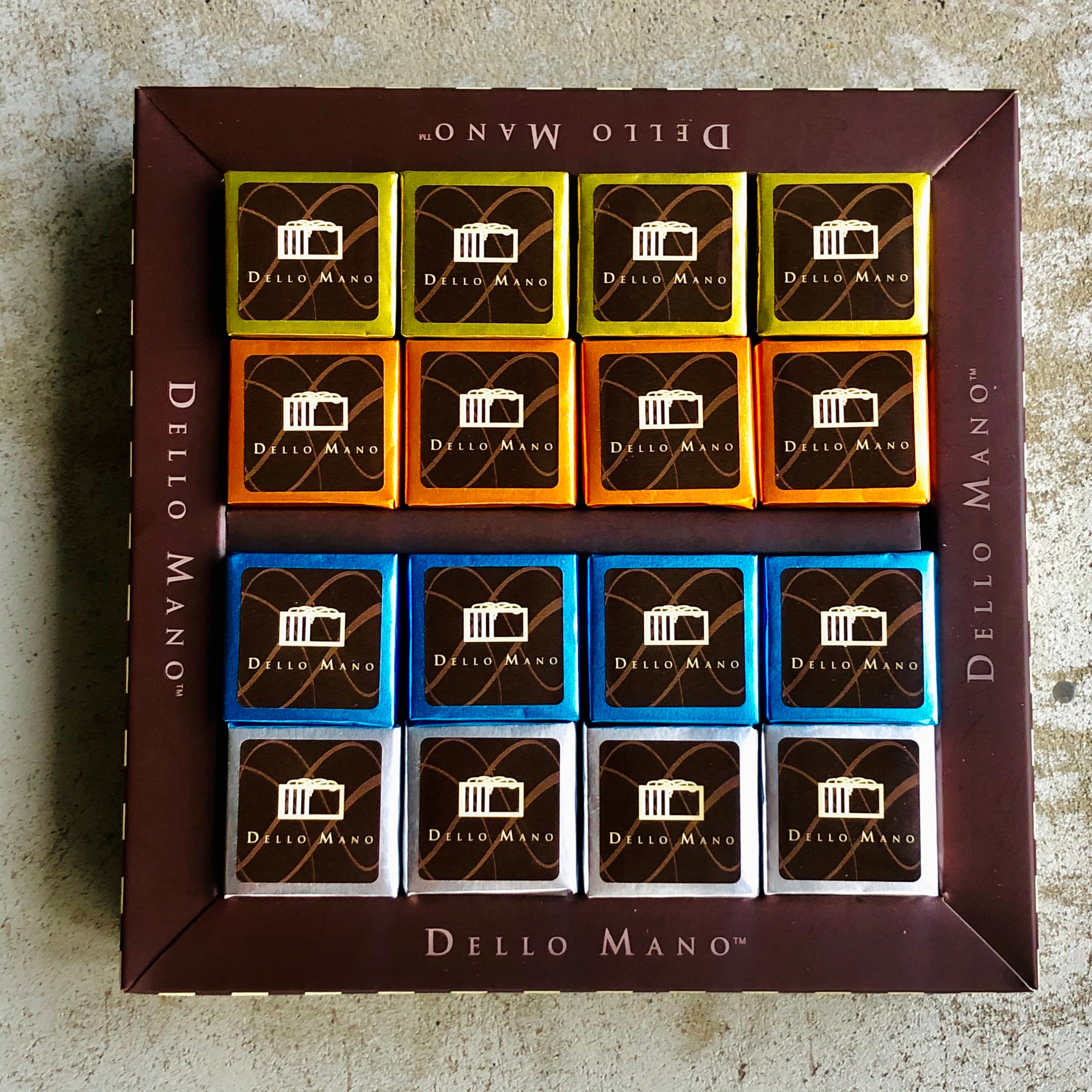Four brownies of each colour gold, orange, blue and silver nestled into a chocolate gift box that has the words Dello Mano printed