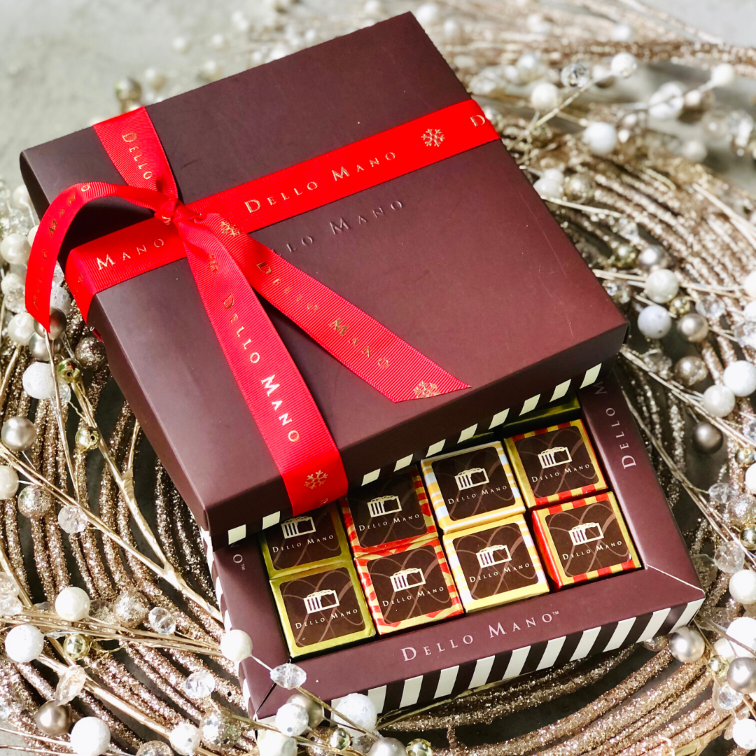 A Sampler gift box of mixed Christmas Brownies in a brownie striped chocolate box. The box has the words Dello Mano.  Another chocolate gift box sits on top with red Christmas themed ribbon and also says Dello Mano