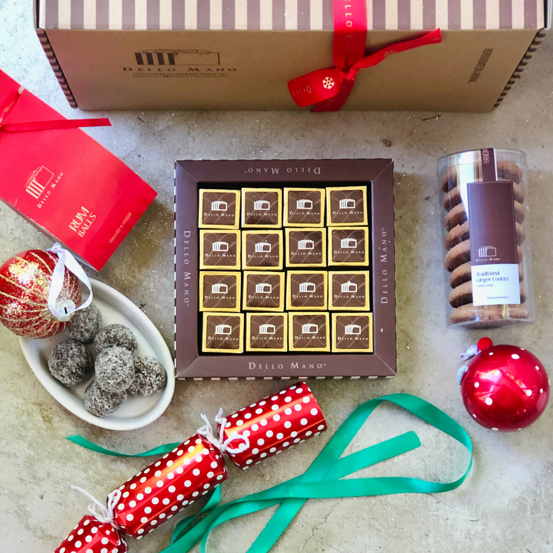 A flat lay image of hamper box, striped gift box of foiled Dello Mano gold brownies , a white bowl of rum balls and a laid flat tube of ginger cookies. A festive Christmas cracker sits at the front with green ribbon and red Christmas bauble. 