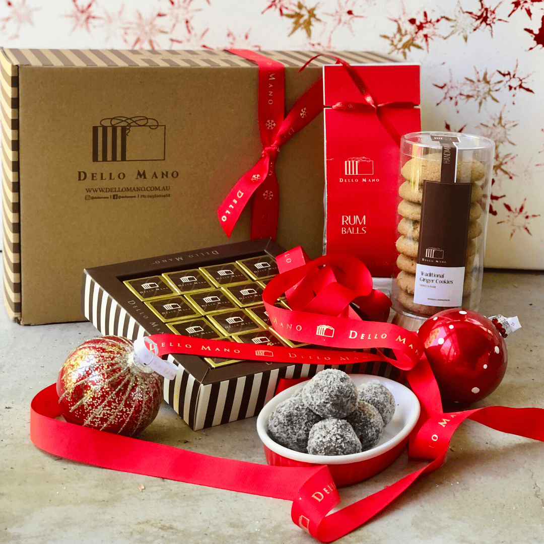 Dello Mano Christmas hamper featuring a kraft gift box with red ribbon, luxury brownies in gold foil, a red box of rum balls, a clear tube of cookies, and a bowl of rum balls, styled with festive red ribbons and ornaments