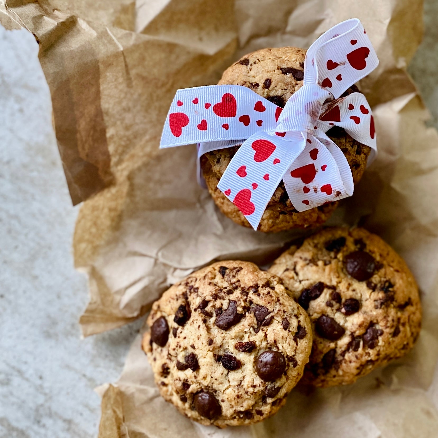 Chocolate Chip Cookies with a stack behind wrapped with white ribbon with red hearts. The cookies are on crumbled brown paper