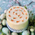The Dello Mano Easter Carrot Cake Gift - a white cake with tiny carrots on top. It sits on a white stand surrounded by green Easter eggs.