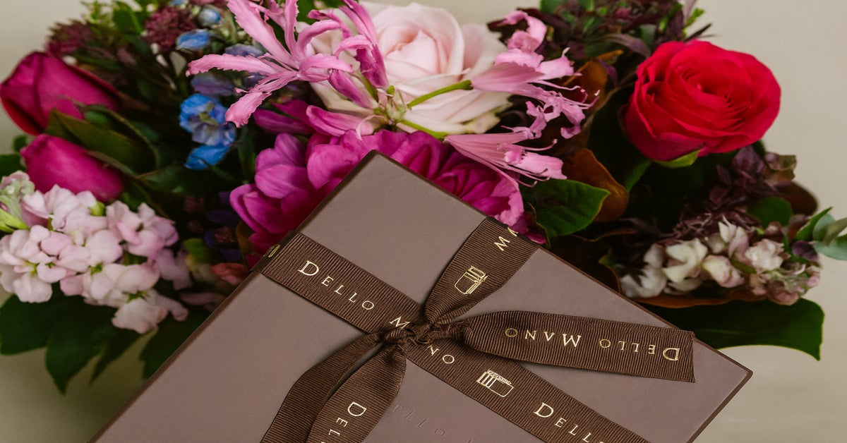 Dello Mano luxury brownie gift box with signature ribbon nestled among vibrant fresh flowers — a thoughtful gift idea that goes beyond flowers.