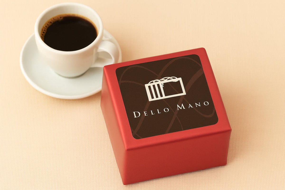 Dello Mano red foiled handmade gluten-friendly brownie cube with signature brown label beside a cup of black coffee on a soft beige surface — a handmade indulgence moment