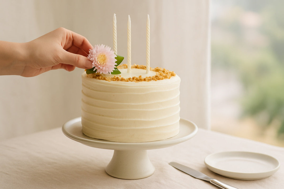Handmade birthday cake in Brisbane on a linen-draped table with soft morning light, a hand gently placing a flower on top, celebrating handmade luxury.