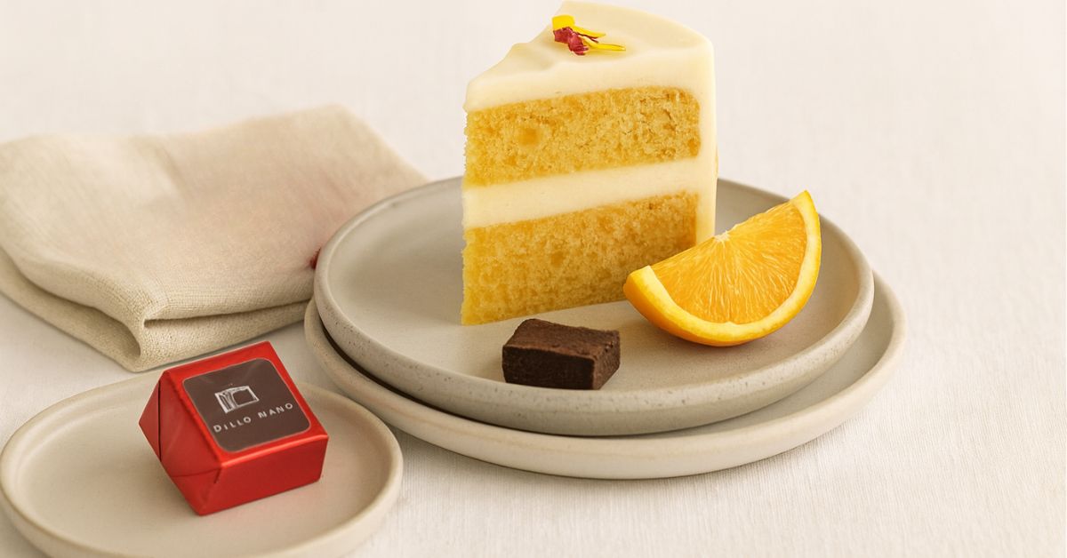 Handmade gluten-friendly orange and almond cake slice with creamy frosting, served beside a Dello Mano gluten-friendly brownie cube wrapped in red foil. Presented on handmade ceramic plates with a linen napkin, evoking calm handmade luxury.