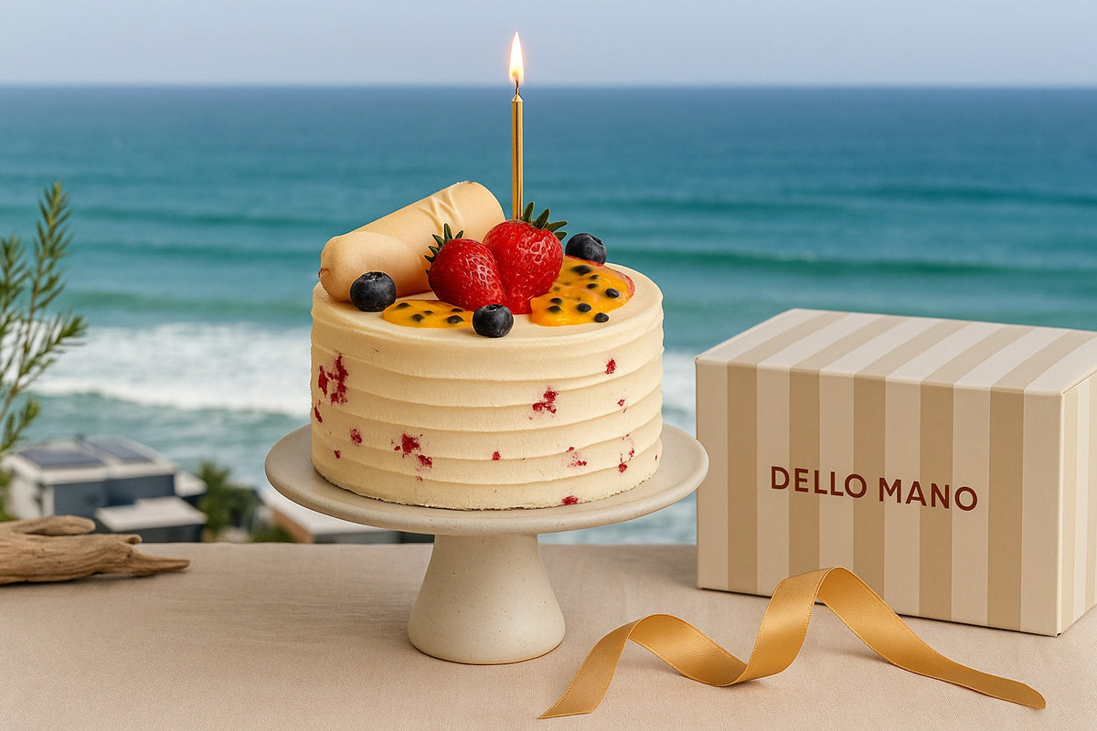 Handmade birthday cake with fresh fruit and gold candle on a table overlooking the Sunshine Coast ocean, with Dello Mano striped box and ribbon beside it.