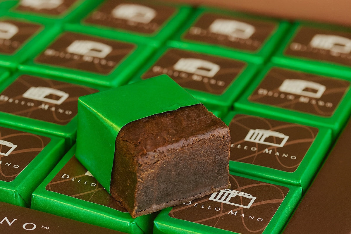 Close-up of Dello Mano vegan brownies wrapped in green foil with the logo visible, featuring one unwrapped handmade brownie showing its rich, fudgy chocolate texture on a striped gift box.
