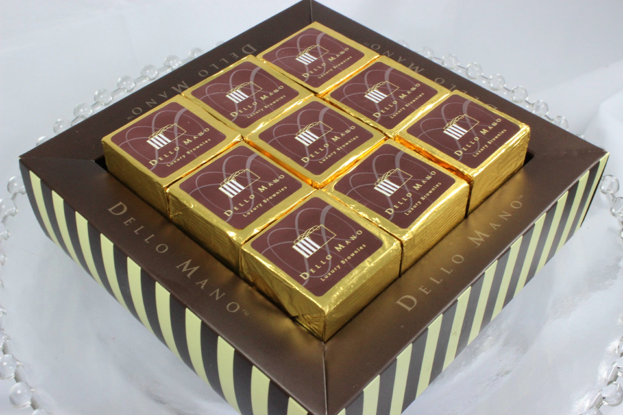 Nine-piece box of Dello Mano luxury brownies with gold wrapping in elegant striped gift box packaging.