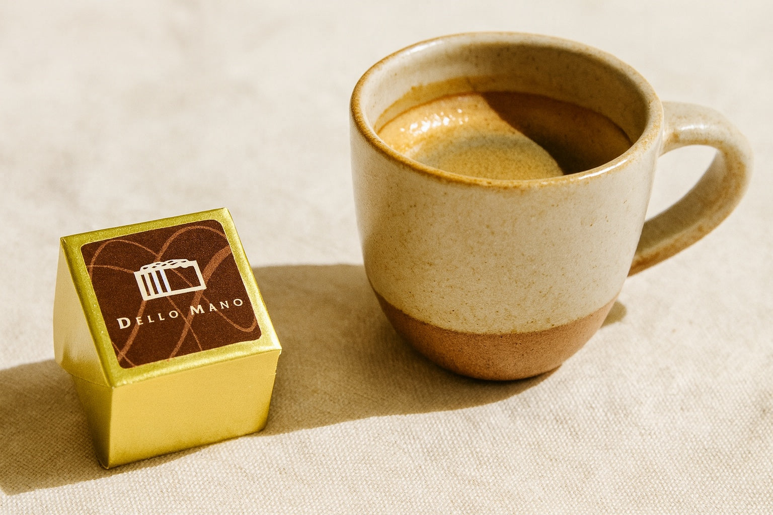 Classic Luxury Brownie Cube by Dello Mano, wrapped in gold foil with branded label, placed beside a handmade ceramic espresso cup on a beige linen background.