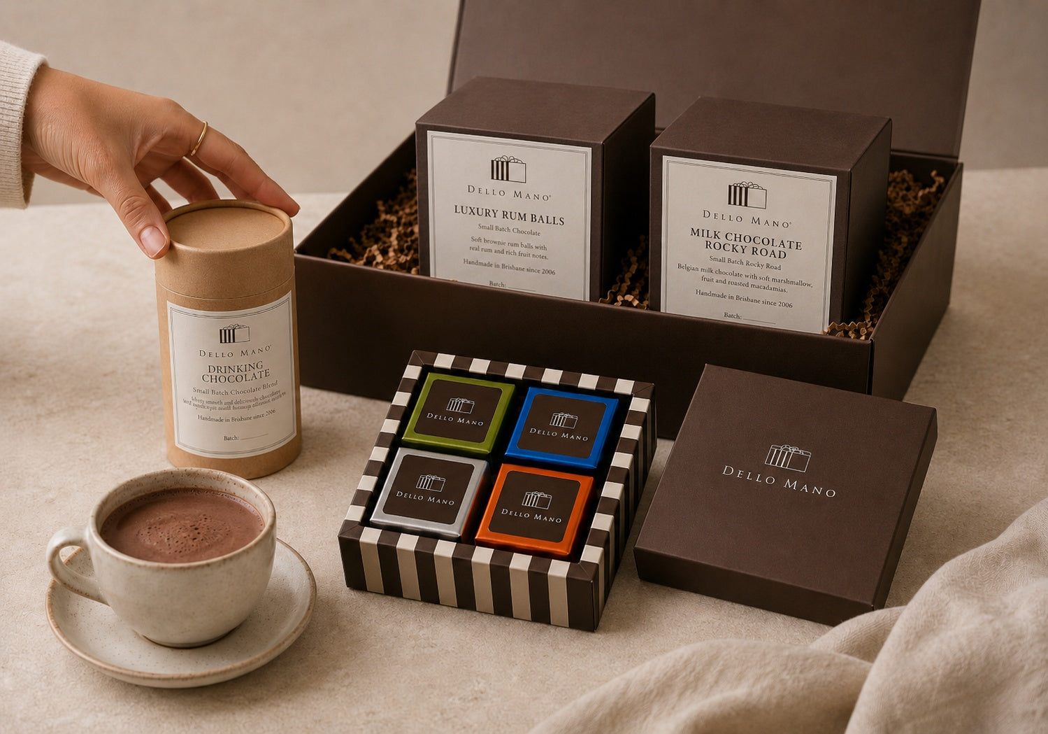 Dello Mano Chocolate Discovery Hamper products arranged on a warm neutral surface with a prepared cup of Drinking Chocolate, a closed four-piece brownie gift box, Luxury Rum Balls and Milk Chocolate Rocky Road in square boxes, and one hand selecting the Drinking Chocolate.