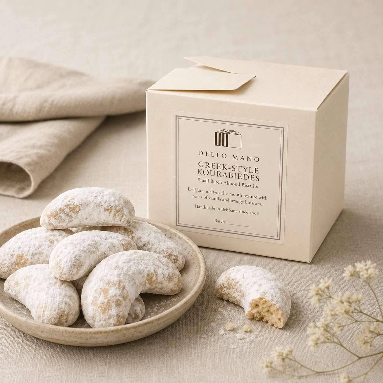 Dello Mano Greek-Style Kourabiedes almond biscuits dusted in icing sugar, styled on a ceramic plate beside a cream gift box on a soft linen background.