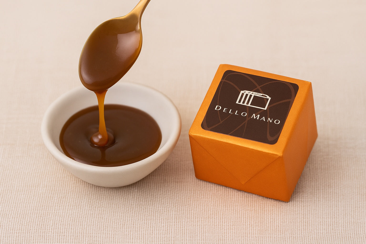 A spoon dripping caramel into a small bowl beside a Dello Mano orange foiled Salted Caramel brownie,
