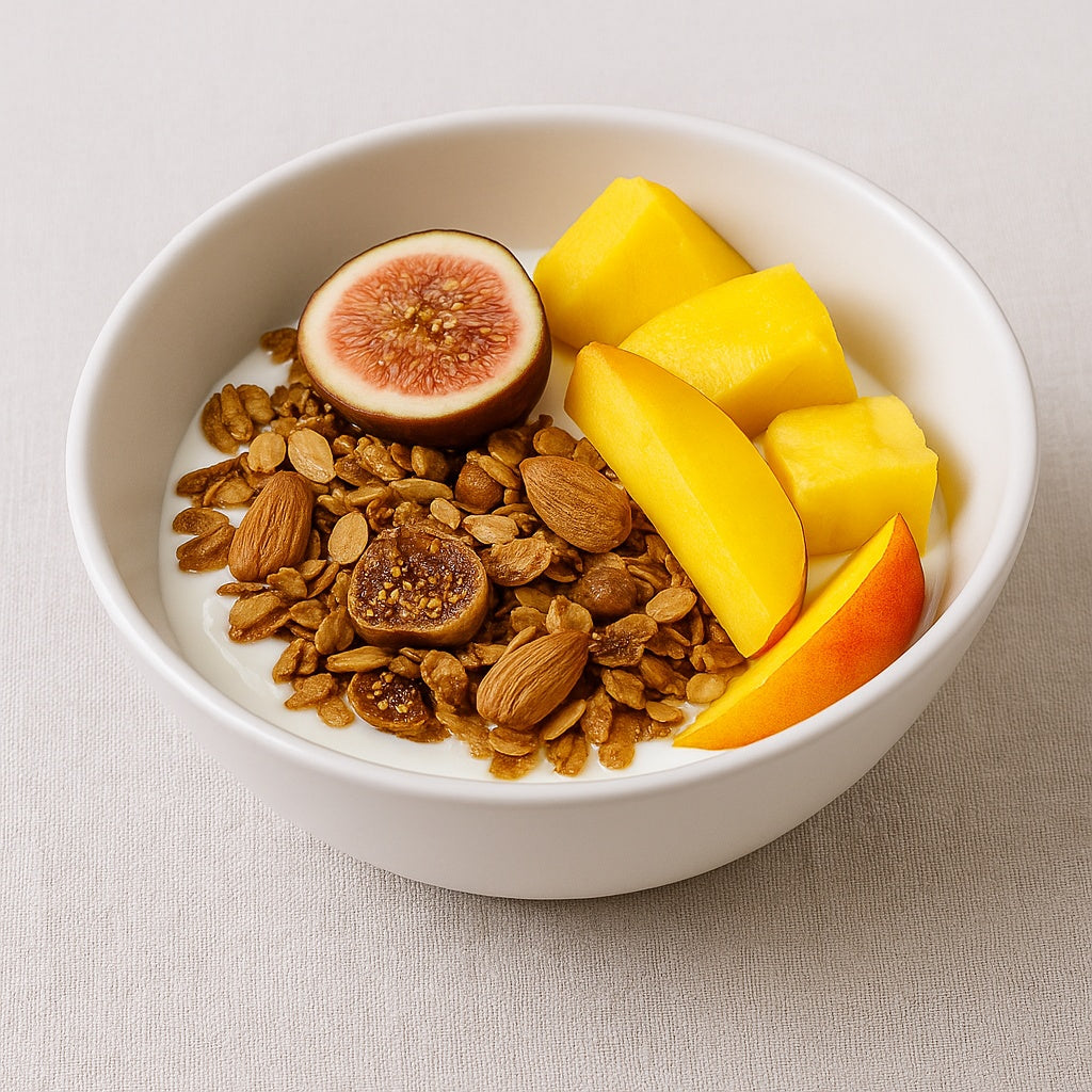 White ceramic bowl of Dello Mano Fig & Almond Granola with yoghurt, topped with dried figs, whole almonds, mango, and peach slices, styled on a linen cloth background in calm natural light