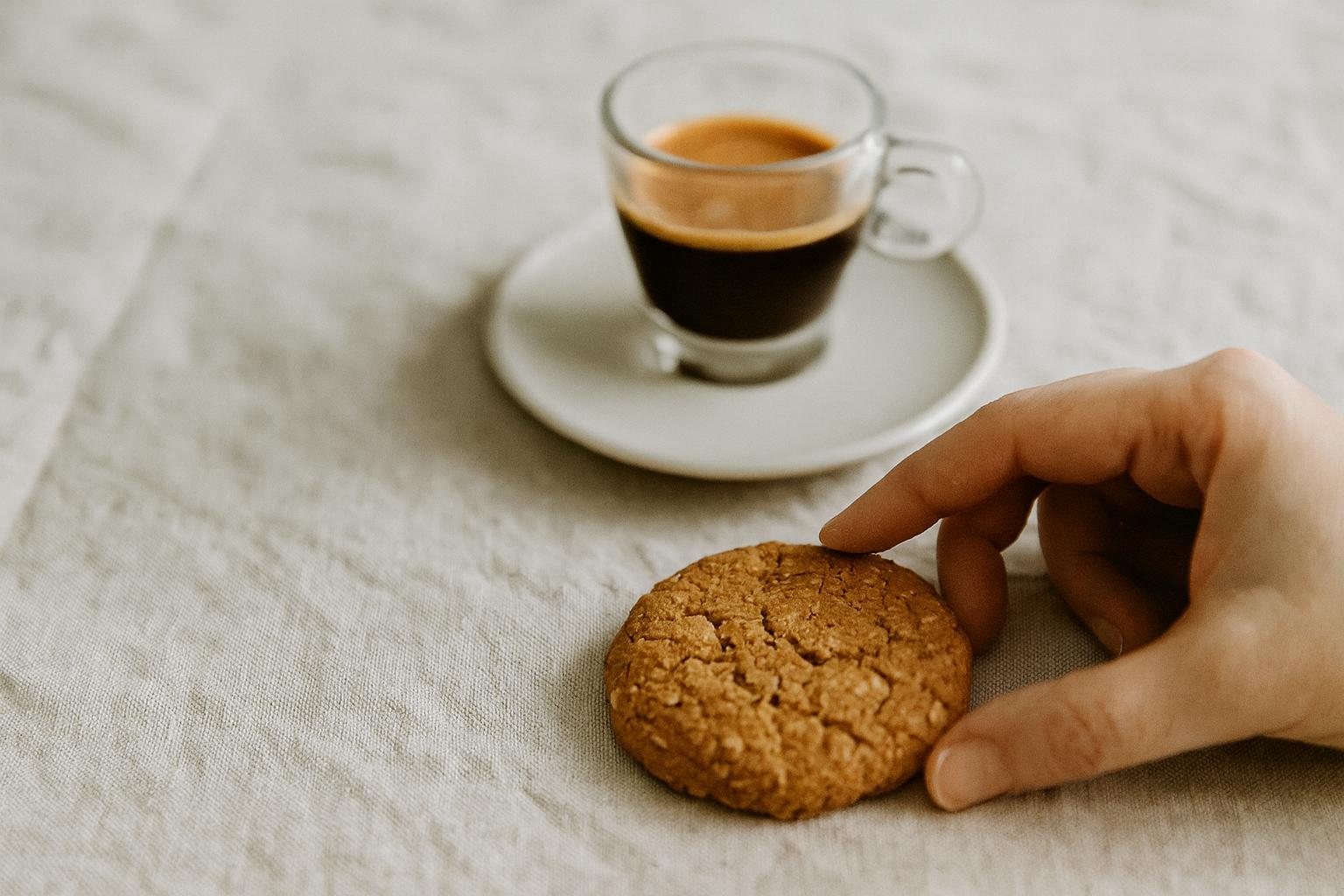 Hand reaching for a handmade caramelised oat and coconut cookie on a natural linen backdrop, with an espresso in a glass cup in the background.