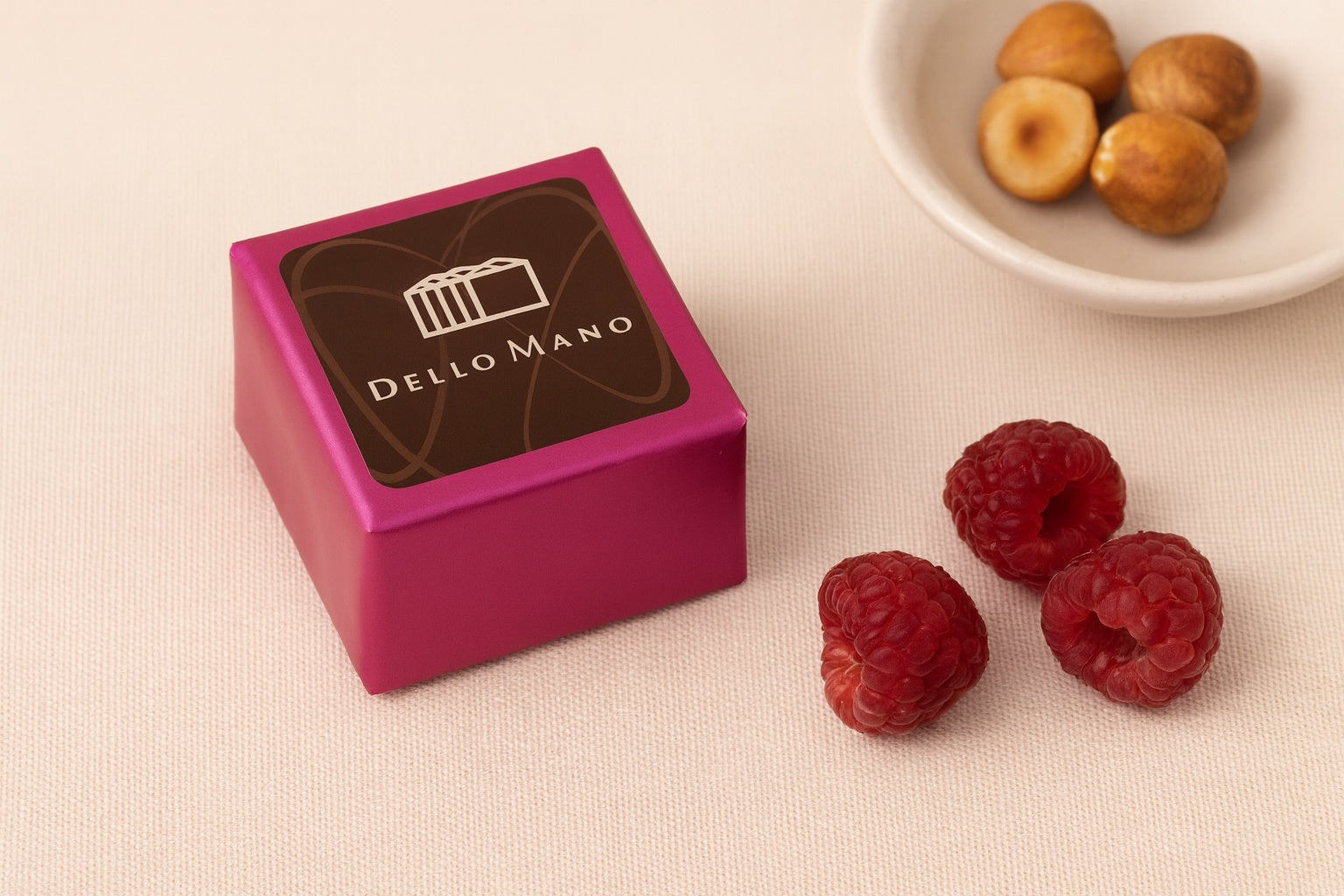 Dello Mano Raspberry Hazelnut Brownie Cube wrapped in pink foil, styled on a beige linen background with fresh raspberries and roasted hazelnuts.