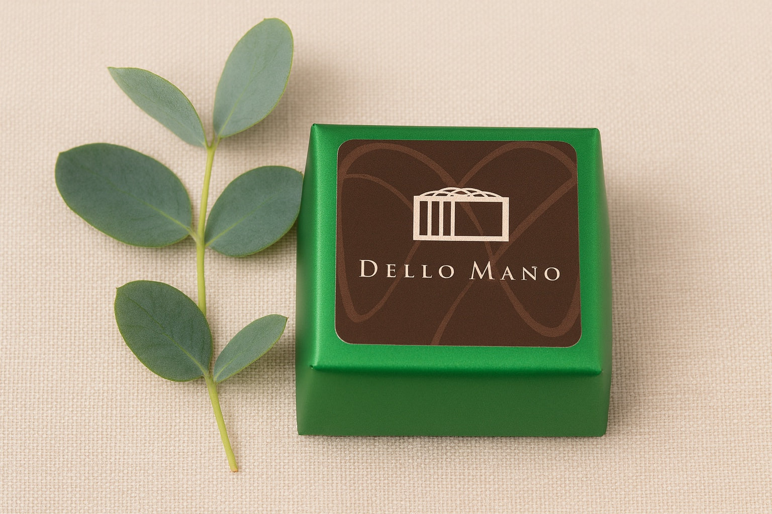 Dello Mano Vegan Brownie Cube wrapped in bright green foil with branded label, styled on a calm beige linen background with a delicate eucalyptus sprig for a subtle, plant-based accent