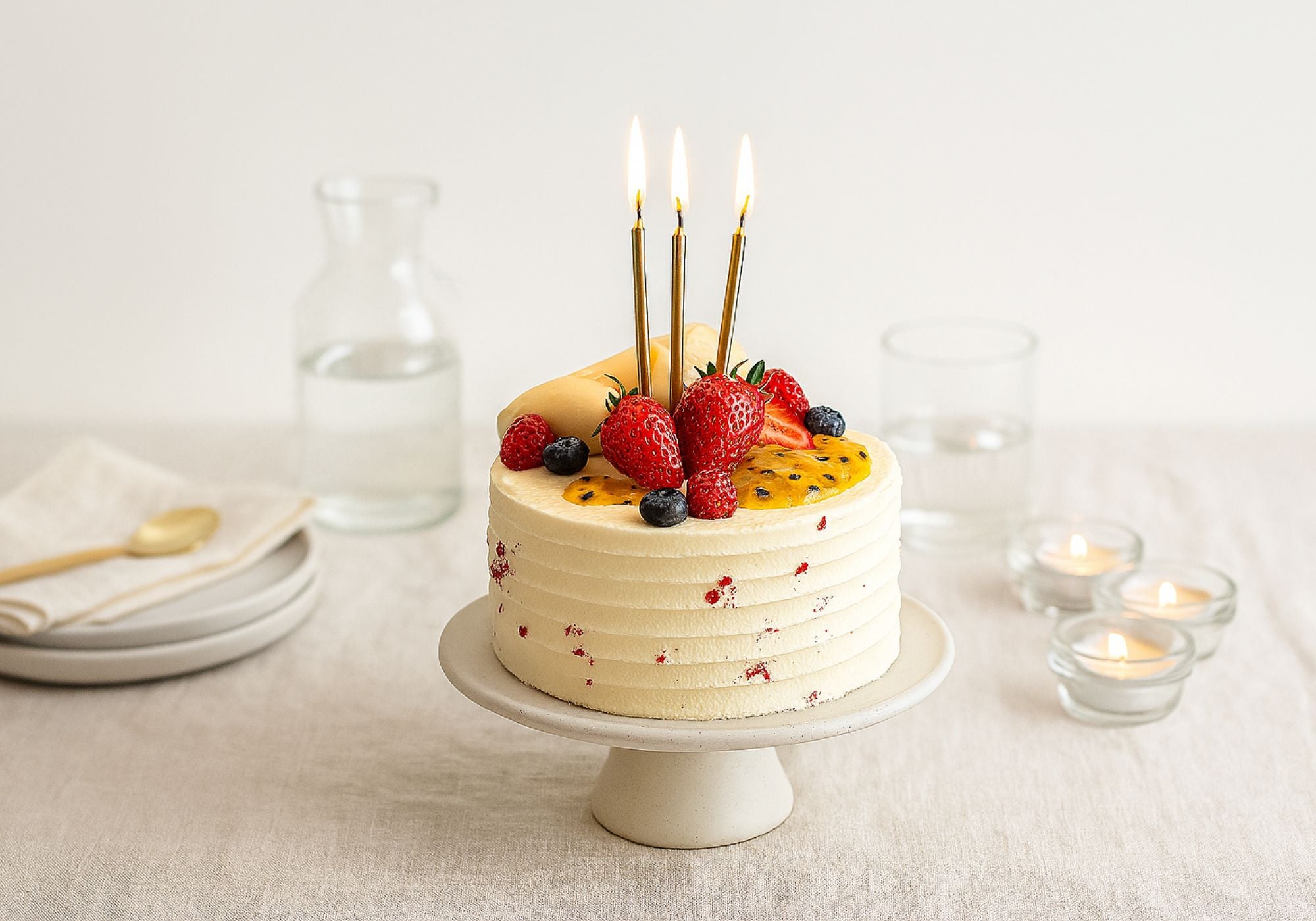 White chocolate birthday cake with fruit toppings and lit candles on a white tablecloth.