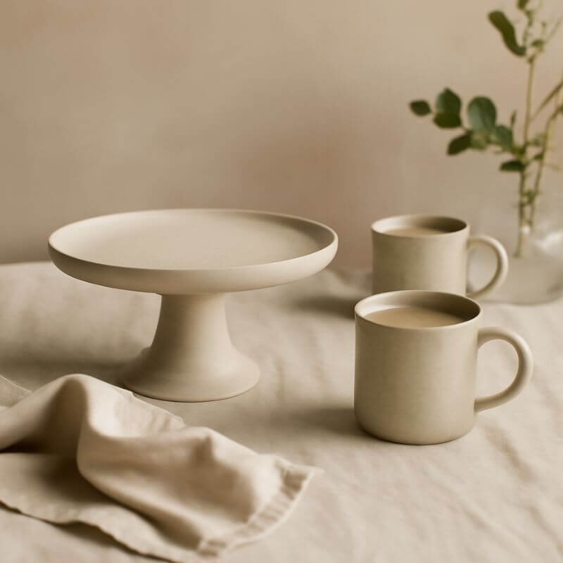 Two ceramic mugs and a cake stand on a neutral surface with a soft background