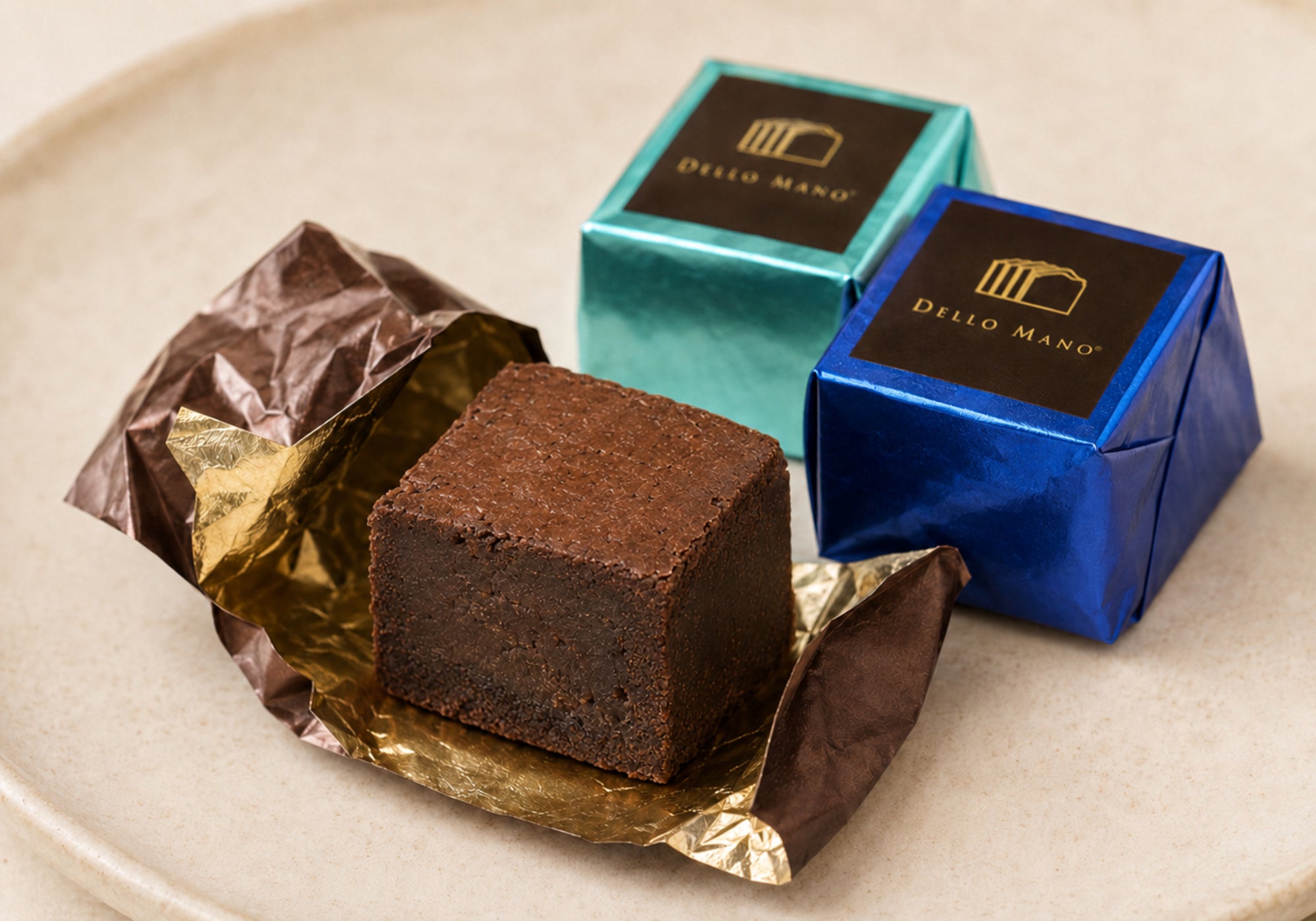 Unwrapped Dello Mano Belgian chocolate brownie showing its dense fudgy centre, with two individually foil-wrapped brownies behind it.