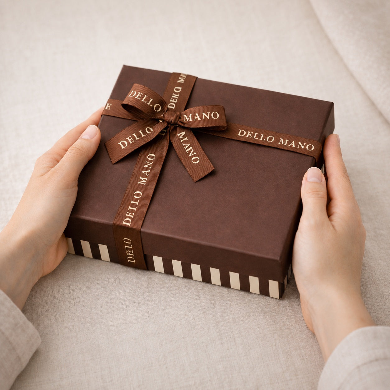 Dello Mano brownie gift box tied with branded brown ribbon, held in two hands against a soft neutral linen background.